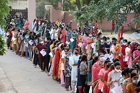 A long line of women wearing colorful clothing and face masks wait outside, each holding papers, alongside a pathway lined with trees.