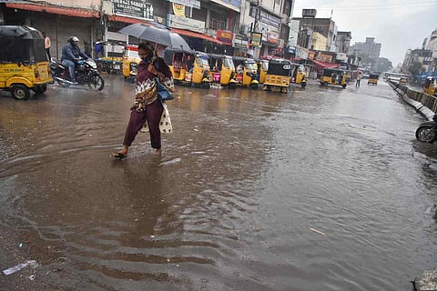 A woman walks barefoot through a waterlogged street holding an umbrella during heavy rainfall in a busy market area, with auto-rickshaws lined up and motorcycles navigating the flooded road. 