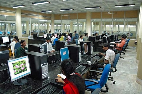 A large hall in which several employees are seated on desks arranged in rows. Each of them is facing a desktop computer