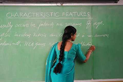 A woman in a teal saree writing on a green chalkboard in a classroom. The board is filled with handwritten text under the heading 'CHARACTERISTIC FEATURES.' She is facing the board with her back partially to the camera, and her long braid hangs down her back.