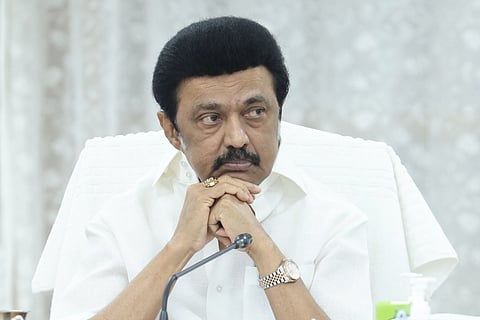 Tamil Nadu Chief Minister MK Stalin at a mic, dressed in a white shirt and seated before a white curtain.