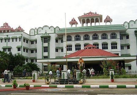 A file image of the Madurai Bench of the Madras High Court. The building is painted white and green. There is a red terracotta roof over the front portico. There is also a garlanded statue of MK Gandhi in front of the building. Several people can be seen walking about. There is a garden in front of the building. 