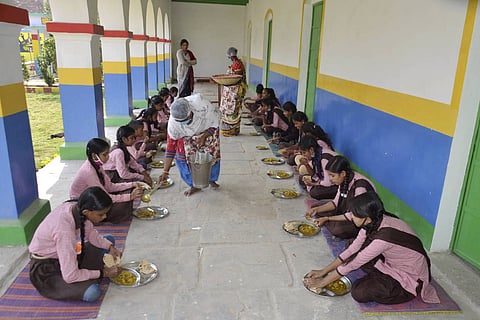A photograph shows female students in pink uniforms sitting on mats in an outdoor corridor. Staff members are serving food from large buckets into the students' metal plates.