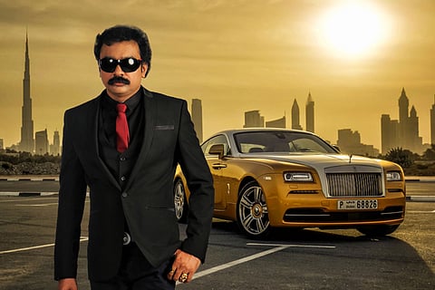 A picture of antique collector Monson Mavunkal in a black suit and red tie, a yellow luxury car can be seen behind him