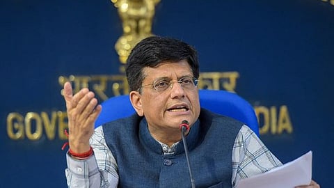 Piyush Goyal in a white shirt with blue check-lines. He is also wearing a blue sleeveless jacket. He is seated on a blue chair and is addressing an audience, with a set of papers in his left hand.