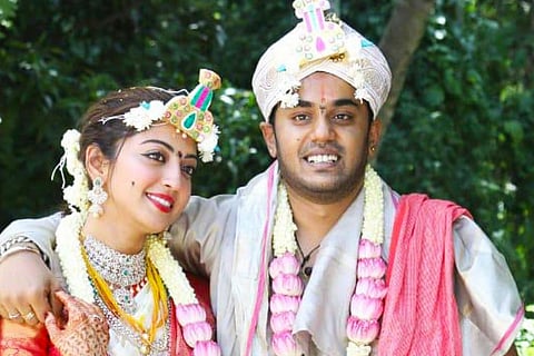 Pranitha is seen wearing a white saree with red borders and Her husband Nitin, seen with his arm around her shoulder and is wearing a festive dhoti in the image.
