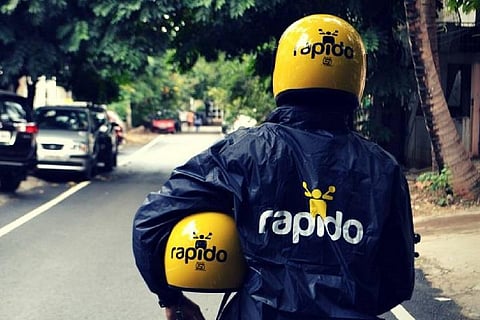 A backside image of a person wearing a jacket and helmet with Rapido logo, holding another helmet with the logo 