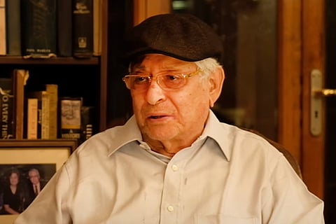 Soli Sorabjee wears a cap and looks to the left with a showcase of books behind him 