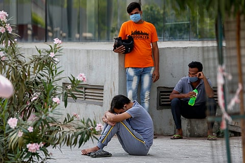 Three young men, one sits with his head bowed, one stands behind in an orange shirt wearing a mask, and a third sits on a platform holding a bottle of water