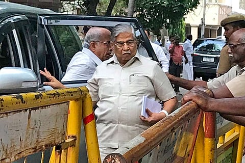 Shanmuganathan is seen walking in a creme safari suit near a police barricade, a car parked next to him