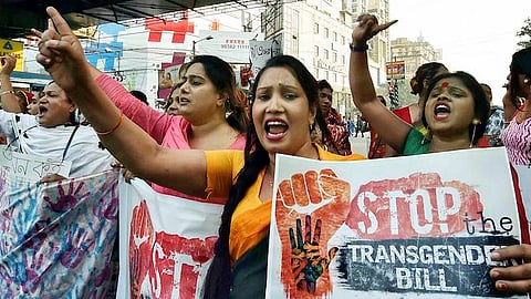 A person in a black saree and yellow blouse raising a slogan and holding a placard that says 'stop the transgender bill'. Other protesters stand around them