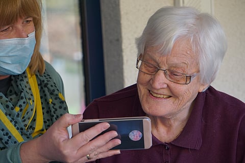 Elderly lady smiles at phone image held by masked woman