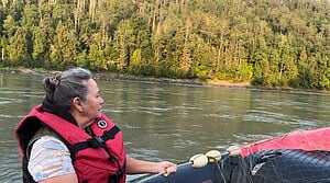 Hereditary Chief Darlene Simpson wears a life jacket while traveling by boat on a calm river lined with dense forest in the "traditional territory" of the Tsetsaut Skii km Lax Ha Nation.