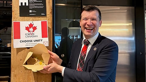 Calgary Mayor Jeromy Farkas holding a breakfast sandwich in front of a Forever Canadian federalist campaign poster. 